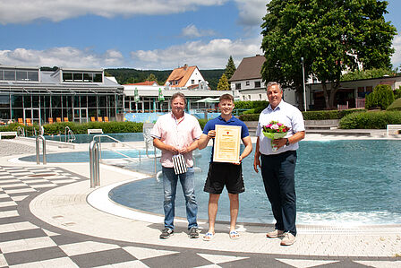 Gratulation zur bestandenen Meisterprüfung! Noah Böhme hat seine Meisterprüfung bestanden. Geschäftsführer Bernd Cranen und Teamleiter Bäder Bernd Kunert gratulieren Herrn Böhme vor dem Nichtschwimmerbecken im Einbecker Freibad und überreichen einen Blumenstrauß und ein Präsent. Herr Böhme hält seine Meisterukunde vor sich.